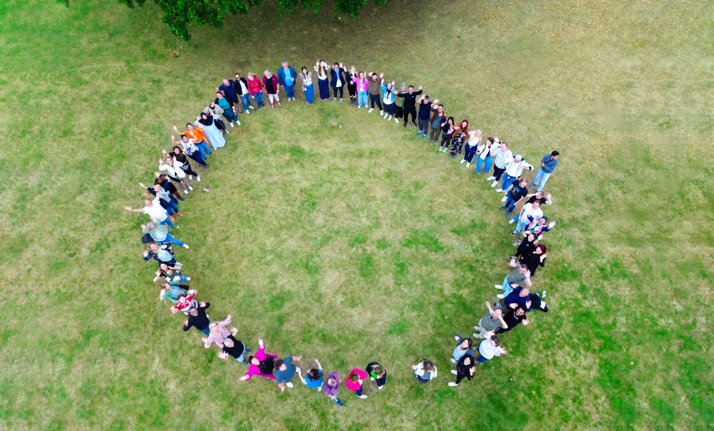 Drohnenaufnahme einer großen Gruppe von Menschen, die sich auf einer Wiese im Kreis aufgestellt haben. Die Personen stehen Schulter an Schulter und blicken nach innen. Im oberen Bildbereich ist ein Teil eines Baumes zu sehen.